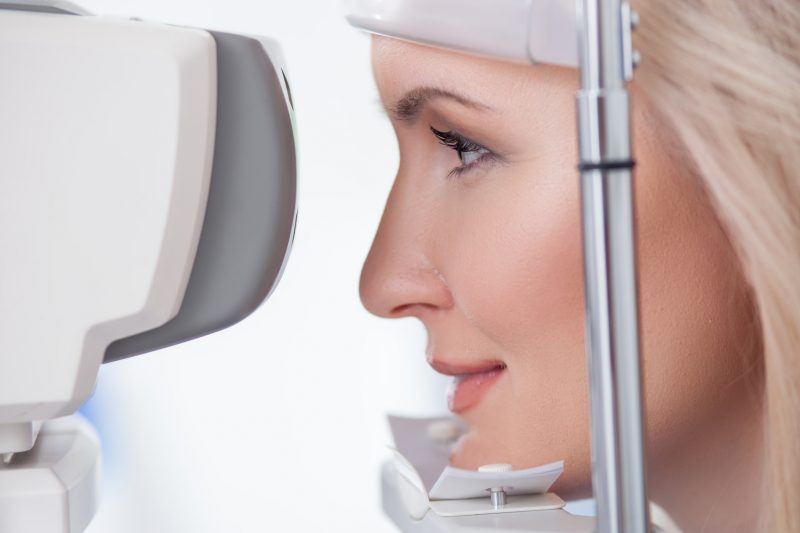 Cheerful blond girl is looking into eye test machine with concentration in oculist lab. She is smiling